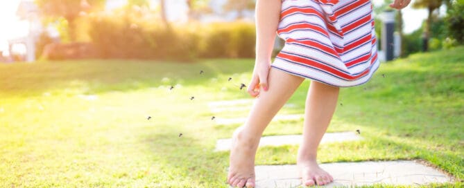 Young girl wearing a tripped dress is scratching her leg as she stands barefoot on outdoors mosquitoes approach her.