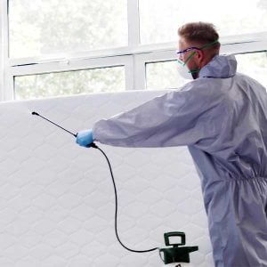Pest control technician in protective gear applying chemical bed bug treatment to a mattress for bed bug treatment in a residential setting.